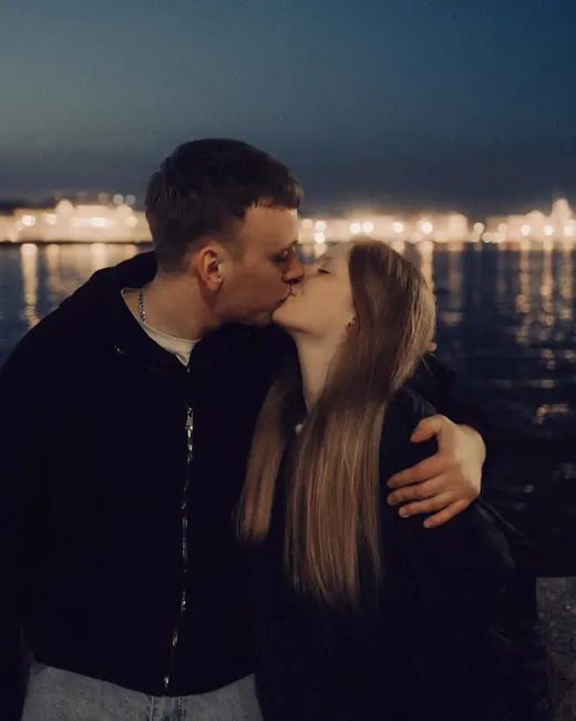 A couple kissing by the water at night, with city lights behind them